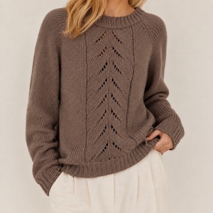 May include: A long-sleeved, taupe-coloured knit jumper featuring a crew neckline and a central vertical panel with an intricate leaf-like eyelet pattern. The garment has ribbed cuffs and a ribbed hem, with raglan sleeves and a solid knit texture.