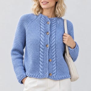 Puede incluir: Un cárdigan de punto azul bígaro con cuello redondo, mangas largas raglán y un patrón de trenzas vertical a lo largo de la tapeta de botones frontal. La prenda se cierra en la parte central delantera con cinco botones redondos claros.