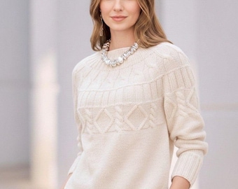 Puede incluir: Suéter de punto de cable color crema con patrones de diamantes y texturizados. El suéter tiene cuello redondo y mangas largas. También se ven un collar y una pulsera plateados.