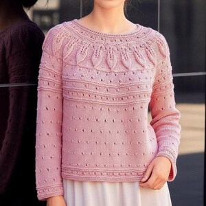 Peut inclure: Un pull rose clair tricoté avec un col rond et des manches longues. Le pull présente un motif de feuilles détaillé sur le haut de la poitrine et des détails ajourés décoratifs. Les manches et le corps ont un motif à pois.