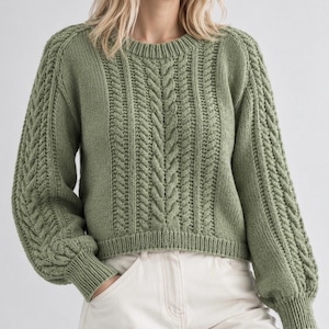 May include: A sage green long-sleeve sweater featuring a prominent vertical cable knit pattern on the front and sleeves, a round crew neckline, and ribbed cuffs and hem. The knit garment is paired with light-colored pants.