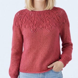 May include: A coral-colored sweater with a round neckline and long sleeves. The yoke features an intricate openwork design. The sweater is made of a soft, knitted material and has ribbed cuffs and hem. The model is wearing blue jeans.
