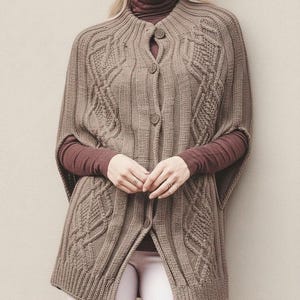 May include: A brown, button-down knitted cape with a cable knit design. The cape has a high collar and short sleeves. The model is wearing a burgundy turtleneck and white pants. The cape is a neutral color, suitable for various outfits.