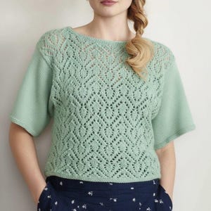 Puede incluir: Top de punto verde claro con cuello redondo y mangas cortas. El top presenta un detallado patrón calado en el pecho y la parte superior de las mangas. La prenda está hecha de un material suave, posiblemente lana.