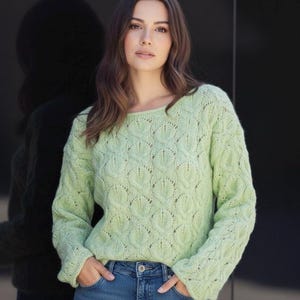 Peut inclure: Un pull vert clair à manches longues avec un motif en forme de feuilles. Le pull a un col rond et est porté avec un jean bleu. La personne porte le pull avec les mains dans les poches.