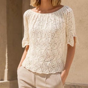 Pode incluir: Uma blusa de manga curta em tom branco sujo com um padrão intrincado de malha aberta repetitivo por toda a peça. O artigo possui um decote largo e arredondado e uma bainha reta, exibindo um design delicado e texturizado.