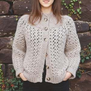 May include: A beige, button-down cardigan sweater with a textured knit pattern. The sweater has a high collar and is fastened with five round, light-colored buttons. The sleeves are long, and the overall style is casual and comfortable.