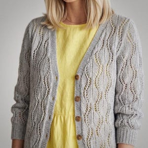 May include: A light grey, open-knit cardigan with a leaf-like pattern. The cardigan has a V-neck, long sleeves, and wooden buttons. The cardigan is worn over a yellow dress.