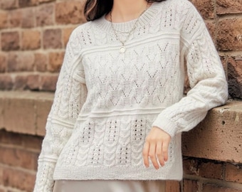 May include: Cream-colored knit sweater with intricate cable and openwork patterns. The long-sleeved sweater features a round neckline and a textured design across the chest and sleeves. The garment is made of a soft, possibly wool, material.