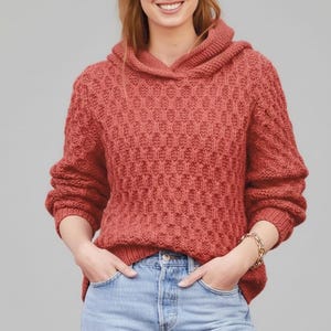 Può includere: Un maglione con cappuccio color rosso mattone con un motivo a maglia strutturato. Il maglione ha maniche lunghe e orlo e polsini a coste. La persona indossa jeans blu e un braccialetto dorato. Lo sfondo è grigio chiaro.