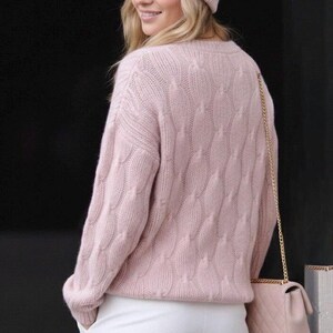 May include: A light pink cable knit sweater with a crew neck. The sweater has a textured pattern and long sleeves. The person is wearing a matching pink beanie and carrying a pink purse with a gold chain strap.