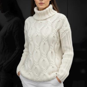 Pode incluir: Camisola de gola alta de malha de cabo creme. A camisola apresenta um padrão texturizado e uma gola alta canelada. As mangas são compridas e o ajuste é folgado. A modelo usa calças brancas. O fundo é preto.