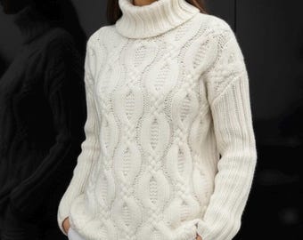 May include: Cream-colored cable-knit turtleneck sweater. The sweater features a textured pattern and a ribbed turtleneck collar. The sleeves are long and the fit is relaxed. The model is wearing white pants. The background is black.