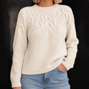 Puede incluir: Un suéter de manga larga color crema con cuello redondo y un prominente patrón de punto trenzado en la parte superior del pecho y los hombros, combinado con jeans azul claro. El suéter tiene cuello, puños y dobladillo acanalados.