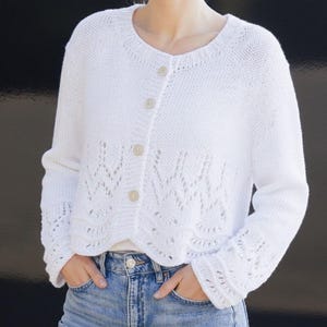May include: A white, button-down cardigan sweater with a round neckline and long sleeves. The sweater features a decorative openwork pattern on the lower front and sleeve cuffs. The buttons are a light beige color. The person is wearing blue jeans.