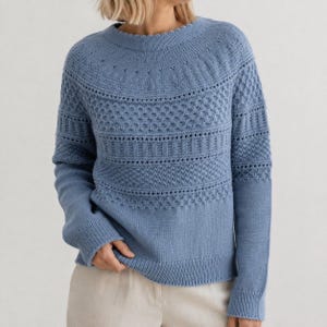 Könnte beinhalten: Ein staubblauer Langarmpullover mit Rundhalsausschnitt und komplexen Strickmustern an Brust und Schultern. Das Kleidungsstück zeigt eine Kombination aus strukturierten Maschenbildern, einschließlich Lochmusterdetails und Rippen.