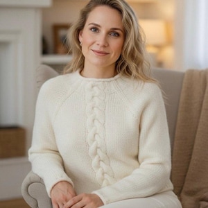 May include: A woman wearing a cream-colored cable-knit sweater. The sweater features a central braided design and a crew neck. She is seated in a light gray armchair, with a neutral-toned blanket draped over the arm. The background is softly blurred.