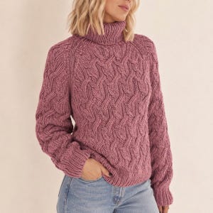 May include: A dusty rose cable-knit sweater featuring a high turtleneck collar, long sleeves with ribbed cuffs, and a textured pattern of interlocking vertical cables throughout the front and sleeves, paired with light blue denim jeans.
