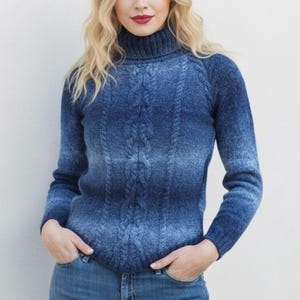 May include: A blue turtleneck jumper with a cable knit pattern. The jumper features a gradient effect, shifting from a darker blue at the top to a lighter blue at the bottom. It has long sleeves and is made of a soft material.