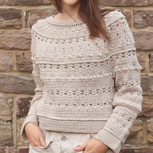 Puede incluir: Un suéter de crochet beige con mangas largas y cuello redondo. El suéter presenta intrincados patrones de puntadas, incluyendo detalles texturizados y calados. La modelo lleva el suéter con pantalones beige.