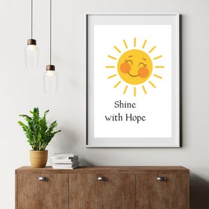 May include: A framed print showing a smiling yellow sun with orange cheeks and yellow rays. The text "Shine with Hope" is below the sun. The print is on a wooden cabinet with a potted plant and hanging lights.