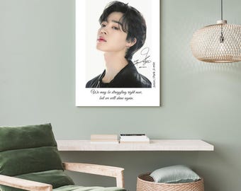 May include: A framed portrait of a person with dark hair and a black jacket. The portrait has a white border and includes the text "We may be struggling right now, but we will shine again." and a signature. The portrait is hanging on a light green wall.