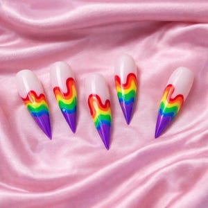 May include: Five long, almond-shaped artificial nails. Each nail features a white base with a rainbow design in red, orange, yellow, green, blue, and purple. The tips are painted purple. The nails are arranged on a pink satin surface.