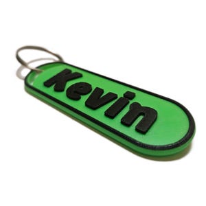 Puede incluir: Un llavero verde con el nombre "Kevin" en letras negras en relieve. El llavero tiene un borde negro y un llavero plateado. El diseño es simple y moderno, adecuado para uso personal.