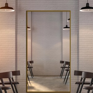 May include: A large rectangular mirror with a gold frame hangs on a white brick wall. The mirror reflects a row of chairs and the opposite wall. Black pendant lights hang from the ceiling.
