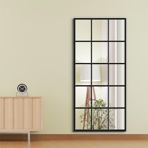 Puede incluir: Un espejo rectangular con marco negro y un patrón de cuadrícula, que se asemeja a una ventana. El espejo refleja una habitación con una lámpara, una planta y cortinas. El espejo está montado en una pared de color claro sobre un suelo de madera. Un pequeño reloj está sobre un mueble de madera.