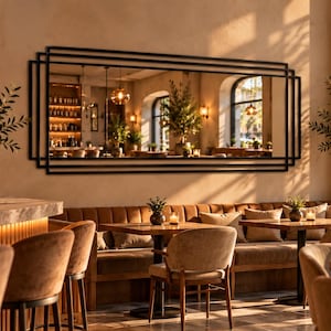 Op de afbeelding: Een grote rechthoekige wandspiegel met een zwart, meerlaags frame met concentrische rechthoekige lijnen. De spiegel is bevestigd aan een beige muur boven een lange, bruin gestoffeerde bank in een restaurantomgeving met warme verlichting.