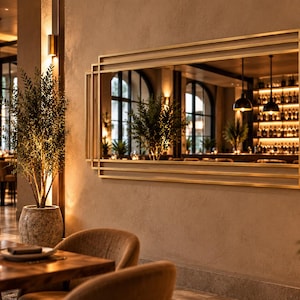 May include: A large rectangular wall mirror featuring a layered gold metal frame with geometric lines. The mirror reflects a restaurant interior with warm lighting, a bar area with shelves of bottles, and green plants, creating a layered visual effect.