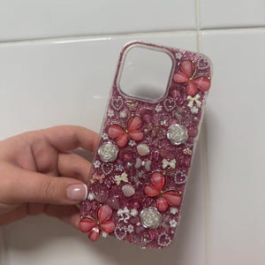 May include: A pink glitter phone case adorned with pink butterflies, white roses, bows, and small beads. The case features a clear camera aperture and is embellished with various decorative elements.