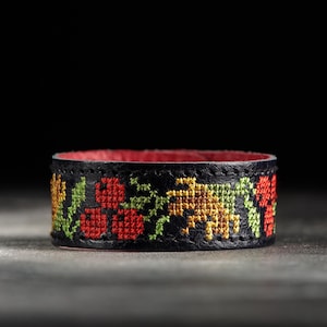 May include: A black leather bracelet with a cross-stitched floral design in red, yellow, and green. The interior of the bracelet is red. The bracelet is a cuff style.