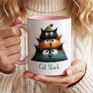 May include: A white ceramic mug with a pink handle and interior, featuring a graphic of three stacked cats in black, orange, and dark teal. The top cat wears a small green hat. The text Cat Stack is printed in black script below the cats.