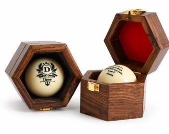 May include: Two hexagonal dark wood boxes with gold-tone latches and red interior lining. Each box contains a cream-colored billiard ball. One ball features a black crest with the letter D and the name Drew. The other ball has black text.