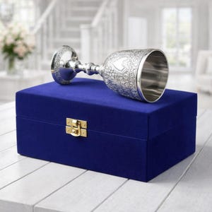May include: A silver goblet with ornate floral detailing rests on a closed, deep blue velvet box with a gold-colored clasp. The goblet's stem and base are also intricately designed. The box sits on a white wooden surface.
