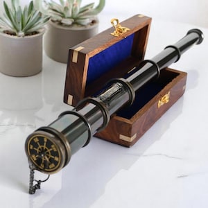 Peut inclure: Un télescope noir et laiton avec un motif de boussole, posé sur une boîte en bois avec un intérieur en velours bleu. La boîte est ouverte, révélant le télescope. Objet décoratif.