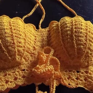May include: A handmade, yellow crochet top with a scalloped edge and halter straps. The top features a detailed, textured pattern and a tie closure at the center. This garment is a unique, handcrafted piece.