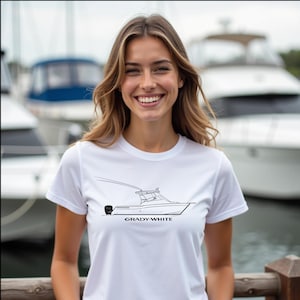 Puede incluir: Camiseta blanca con un dibujo lineal negro de un barco Grady-White. La camiseta tiene cuello redondo y mangas cortas. El texto "GRADY-WHITE" está impreso debajo de la ilustración del barco. El fondo muestra barcos y agua.