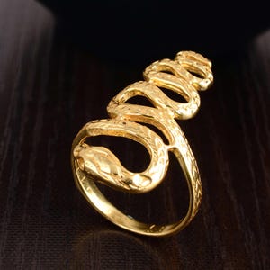 Puede incluir: Un anillo dorado con forma de serpiente, enrollado alrededor del dedo. La cabeza de la serpiente está detallada con escamas texturizadas y una boca ligeramente abierta. El anillo tiene una superficie texturizada y está sobre un fondo oscuro.