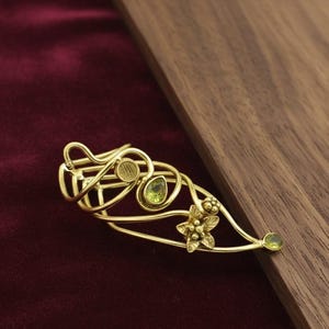 May include: A gold-toned brooch with an intricate, swirling design. The brooch features a floral element and several small, oval-shaped green gemstones. The piece is set against a dark red background and a wooden surface.