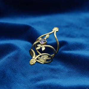 May include: A gold-colored ring with a rectangular purple gemstone. The ring features a floral design with leaves and a decorative top. The ring is set against a blue background.
