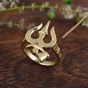 Puede incluir: Un anillo dorado con un diseño de tridente. El anillo presenta un símbolo de tridente de tres puntas, un elemento central en forma de campana y detalles decorativos a lo largo de la banda. El anillo está sobre una superficie de madera oscura.