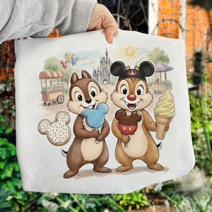 May include: A white tote bag featuring Chip and Dale enjoying treats in front of a Disney park scene. Chip holds a blue Mickey Mouse-shaped ice pop, while Dale has a Mickey Mouse-shaped caramel apple and a soft-serve ice cream cone.