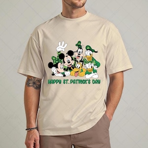 Puede incluir: Camiseta beige con un diseño de Mickey y Minnie Mouse, Goofy, Donald Duck y Pluto, todos vestidos de verde para el Día de San Patricio. El texto "HAPPY ST. PATRICK'S DAY" está impreso en verde debajo.