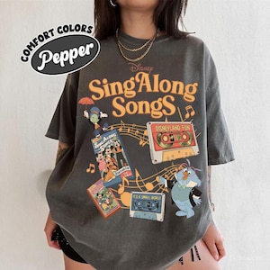 May include: Dark grey t-shirt featuring the text "Disney Sing Along Songs" in orange, with cartoon characters and cassette tape graphics. The shirt also has the text "Disneyland Fun" and "It's a Small World". The shirt is labeled "Comfort Colors Pepper".