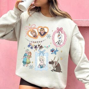 Könnte beinhalten: Ein hellbeige Sweatshirt mit farbenfrohen Illustrationen von Charakteren aus dem Animationsfilm "Die Eiskönigin". Das Design umfasst gerahmte Porträts von Anna, Elsa, Kristoff und Olaf sowie andere Charaktere und das Wort "FROZEN".