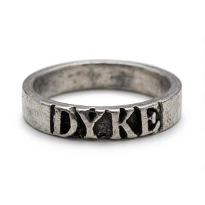 May include: A silver ring with the word "DYKE" engraved on the band. The ring has a worn, textured appearance, and the letters are slightly uneven. The ring is a simple, yet bold piece of jewellery.