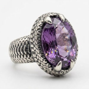 Puede incluir: Un anillo de plata con una gran piedra preciosa morada de corte ovalado engastada en una banda de metal texturizada similar a escamas. La piedra está sujeta por cuatro garras metálicas prominentes y la superficie muestra un patrón detallado.